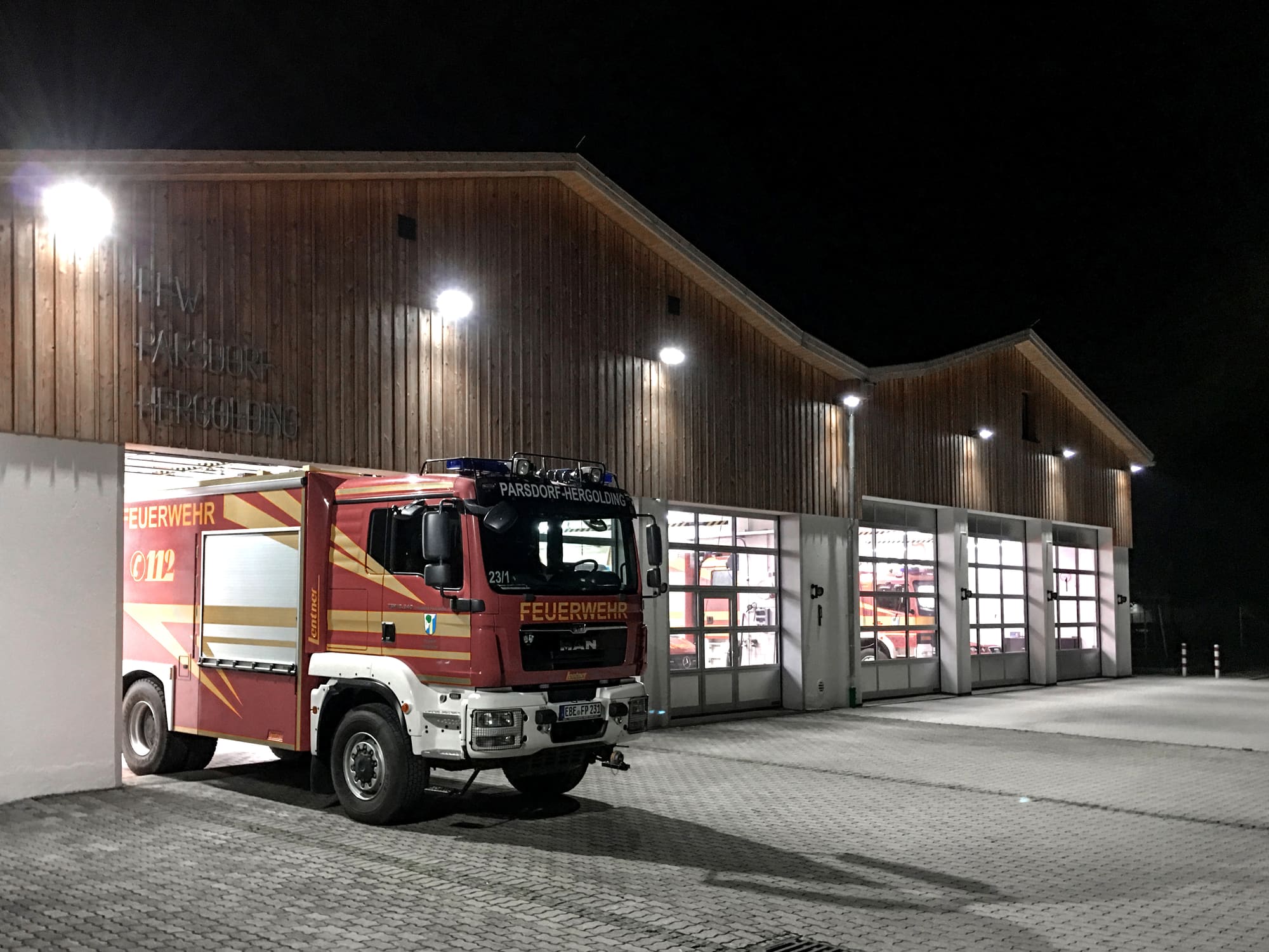 Beleuchtete Fahrzeughalle mit Feuerwehrfahrzeug am Abend – Anbau und Umbau der Feuerwehr Parsdorf